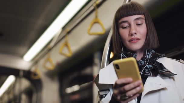 Retrato de uma jovem sorridente em fones de ouvido andando em transportes públicos, ouvir música e navegar em smartphones amarelos. Luzes da cidade fundo. Comunicação, boas notícias, estrada, conceito de viagem . — Vídeo de Stock