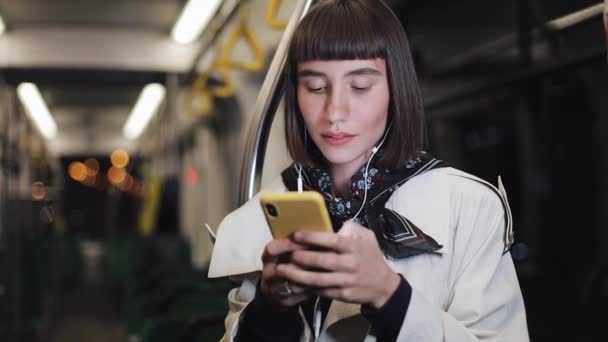 Retrato de una hermosa mujer joven sonriente en auriculares montados en transporte público, escuchar música y navegar en el teléfono inteligente amarillo. Fondo de luces de ciudad. Comunicación, carretera, concepto de viaje . — Vídeo de stock