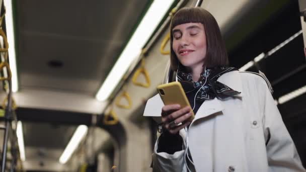 Retrato de una joven sonriente en auriculares montados en transporte público, escuchar música y navegar en un smartphone amarillo. Fondo de luces de ciudad. Comunicación, buenas noticias, carretera, concepto de viaje . — Vídeos de Stock