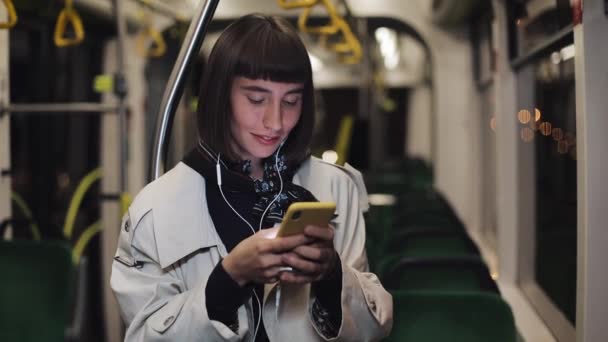Portret pięknej młodej kobiety w słuchawkach jazda w transporcie publicznym, słuchać muzyki i przeglądania na żółtym smartfonie. Tło świateł miejskich. Komunikacja, droga, koncepcja podróży. — Wideo stockowe