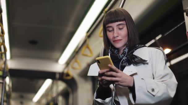 Retrato de una hermosa mujer joven en auriculares montados en transporte público, escuchar música y navegar en el teléfono inteligente amarillo. Fondo de luces de ciudad. Comunicación, carretera, concepto de viaje . — Vídeo de stock