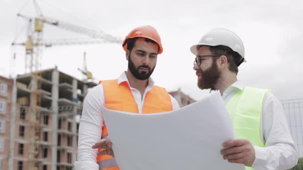 男性建設技術者が高層ビルの建設現場や建築現場で建築家と話し合う。建設図面を手に持つ。. — ストック動画