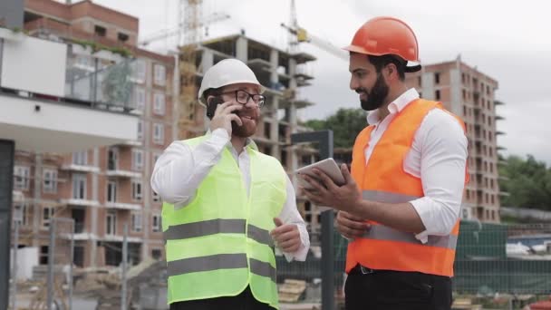 Ingeniero feliz habla en el teléfono móvil en el sitio de construcción y comprueba el trabajo del trabajador. Constructor habla en smartphone. Comprobación de la fase de construcción — Vídeo de stock