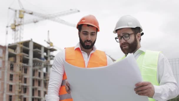 Discussão de engenheiro de construção masculino com arquiteto no canteiro de obras ou canteiro de obras do prédio do arranha-céus. Eles segurando desenhos de construção em suas mãos . — Vídeo de Stock