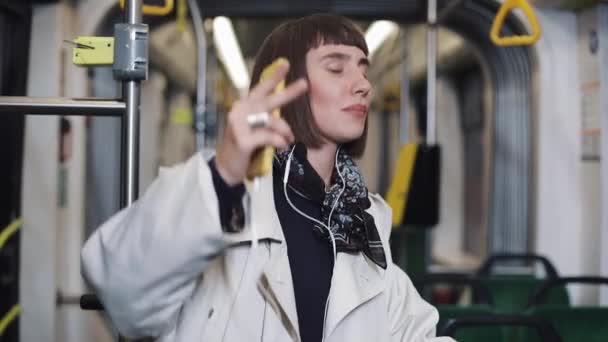 年轻的快乐女人穿着外套与耳机听音乐和有趣的舞蹈在公共交通工具的肖像. — 图库视频影像