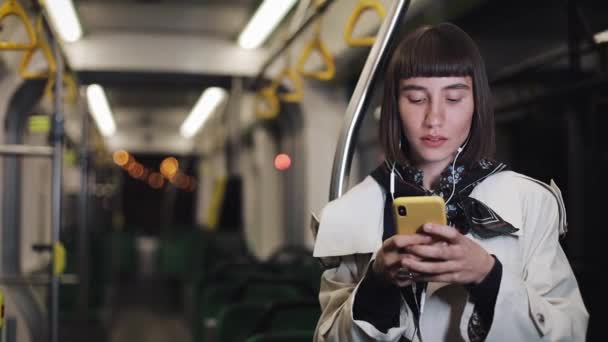 Retrato de bela jovem em fones de ouvido andando em transportes públicos, ouvir música e navegação no smartphone amarelo. Luzes da cidade fundo. Comunicação, estrada, conceito de viagem . — Vídeo de Stock