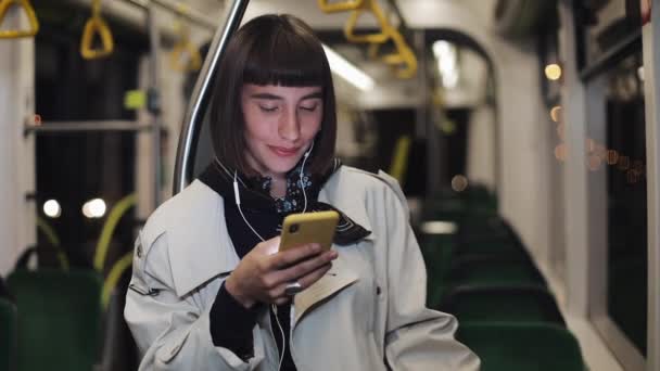 Portrait of beautiful young woman in headphones riding in public transport, listen music and browsing on yellow smartphone. City lights background. Communication, road, travel concept. — Stock Video