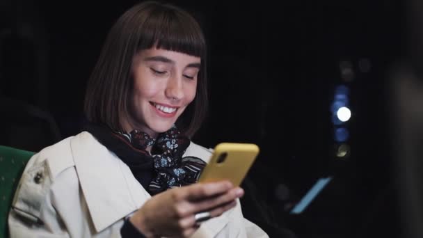 Jolie jeune femme souriante dans les transports en commun à l'aide d'un téléphone portable. Elle envoie des SMS, vérifie les mails, les chats ou les nouvelles en ligne. Fond de lumières de ville . — Video