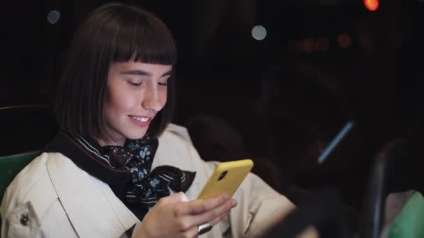 Atractiva joven sonriente en transporte público usando un teléfono móvil. Ella está enviando mensajes de texto, revisando correos electrónicos, chats o las noticias en línea. Ciudad luces fondo . — Vídeo de stock