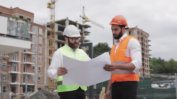 männliche Bauingenieur Diskussion mit Architekt auf der Baustelle oder auf der Baustelle eines Hochhauses. sie halten Konstruktionszeichnungen in ihren Händen.