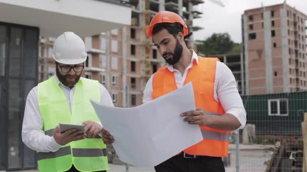 The builder and architect man are discussing the construction plan of the modern business center walking near construction site. They checks the work with a tablet and architectural drafting. — Stock Video