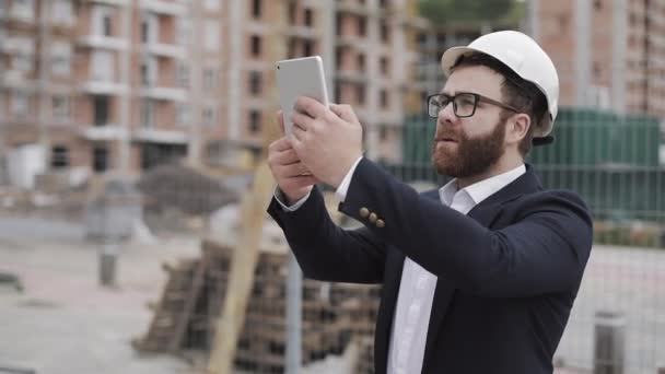 Junger bärtiger Architekt auf der Baustelle beim Fotografieren mit Tablet. er im Businessanzug. — Stockvideo