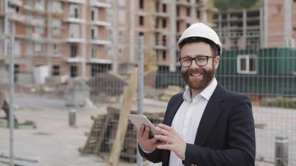 Portret van de jonge zakenman met Tablet op de bouw site glimlachend op zoek naar de camera het dragen van een veiligheidshelm en pak. Ingenieur, architect, bouwer, zakenman. — Stockvideo