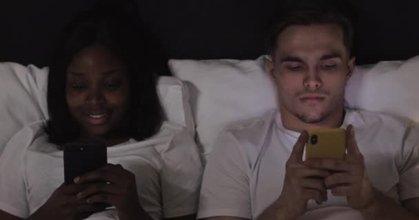 ベッドの中でスマートフォンを持つ幸せな多民族のカップル。人、技術、インターネット、コミュニケーションの概念. — ストック動画