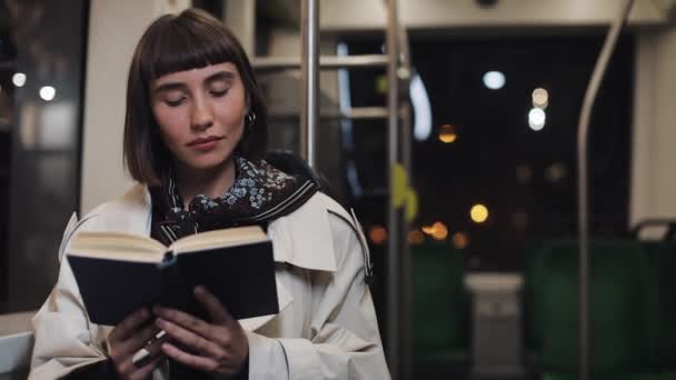 Young woman or passenger reading book sitting in public transport, steadicam shot. Slow motion. City lights background. Commuter, student, knowledge concept. — Stock Video