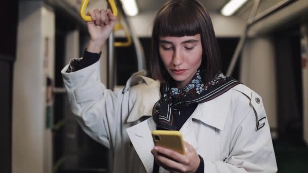 Portret van lachende mooie jonge vrouw in het openbaar vervoer houdt de leuning en browsen op gele smartphone. Stadslichten achtergrond. Communicatie, weg, reis concept. — Stockvideo