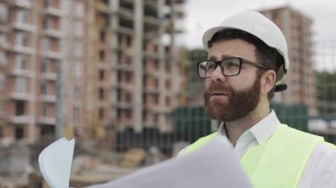 Başarılı bir genç mühendis veya mimar beyaz bir kask giyen portresi, elinde inşaat çizimleri inşaat alanı yakınında duran bakarak.