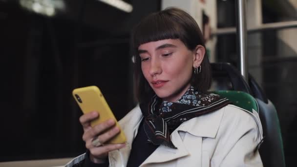 Junge Hipsterfrau im Chat auf Smartphone sitzend in öffentlichen Verkehrsmitteln, Steadicam-Aufnahme. Junge Frau erhält gute Nachrichten per Smartphone. Zeitlupe. Hintergrund Stadtbeleuchtung. — Stockvideo