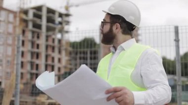 Başarılı bir genç mühendis veya mimar beyaz bir kask giyen portresi, elinde inşaat çizimleri inşaat alanı yakınında duran bakarak.