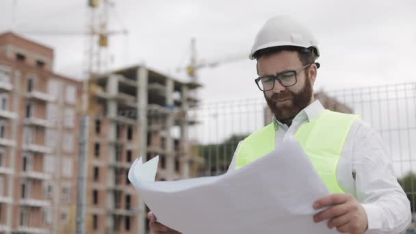 Retrato de un exitoso joven ingeniero o arquitecto con un casco blanco, mirando en los dibujos de construcción en su mano de pie cerca de la obra . — Vídeos de Stock