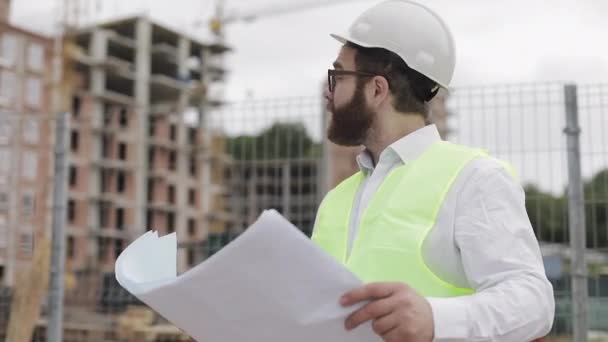 Retrato de um jovem engenheiro ou arquiteto bem sucedido vestindo um capacete branco, olhando nos desenhos de construção em sua mão de pé perto do canteiro de obras . — Vídeo de Stock