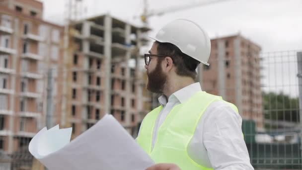Retrato de um jovem engenheiro ou arquiteto bem sucedido vestindo um capacete branco, olhando nos desenhos de construção em sua mão de pé perto do canteiro de obras . — Vídeo de Stock