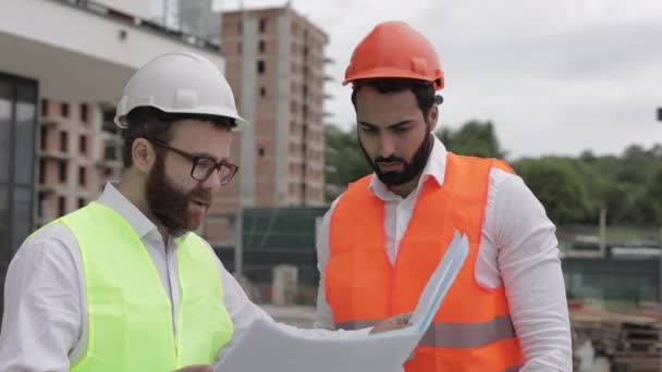 O construtor e arquiteto homem estão discutindo o plano de construção do moderno centro de negócios em pé perto do canteiro de obras. Eles vão para o edifício. . — Vídeo de Stock