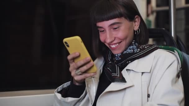 Jovem sorridente atraente no transporte público usando um telefone celular. Ela está enviando mensagens, verificando e-mails, chats ou as notícias on-line. Cidade luzes fundo . — Vídeo de Stock