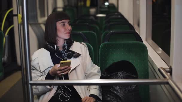 Mujer joven y elegante en los auriculares escuchando música y la navegación en el teléfono móvil a caballo en el transporte público. Ciudad luces fondo . — Vídeo de stock