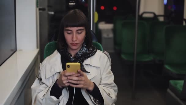 Jeune femme attrayante dans les transports publics en utilisant un smartphone. Elle envoie des SMS, vérifie les mails, les chats ou les nouvelles en ligne. Fond de lumières de ville . — Video