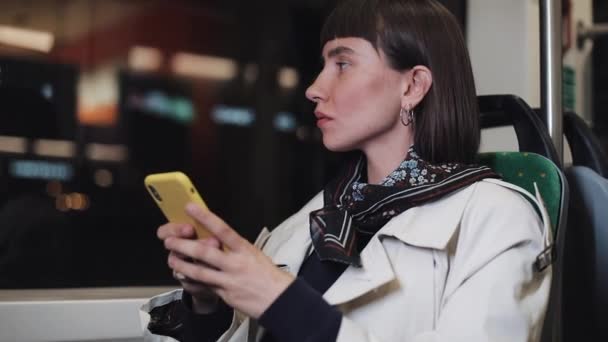 Młoda kobieta przygnębiona siedzi w transporcie publicznym, patrząc z okna pociągu. Trzyma smartfon w ręku. Smutek, Melancholy. Tło świateł miejskich. — Wideo stockowe