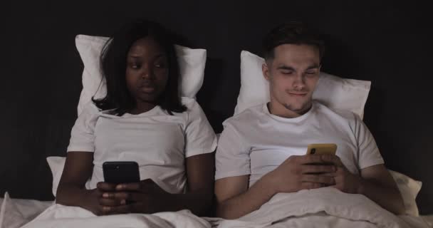 Getrouwd multi-etnisch echtpaar liggend in bed 's nachts en het gebruik van smartphones. Vrouw gluren haar man het openen van sociale media op mobiele telefoon. Spionage, complexe relaties. Geen vertrouwen. — Stockvideo