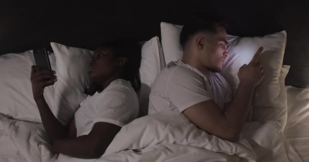 Marié jeune couple multi-ethnique couché dans le lit dos à dos la nuit et tapant ou faisant défiler sur les smartphones. À l'intérieur. Vue du dessus . — Video