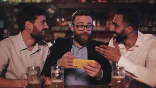 Three old friends watching video on smartphone and drinking draft beer in the sport bar. Friends laughing and discuss the news in beer pub. — Stock Video