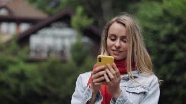Akıllı telefonda uygulamayı kullanan, cep telefonunda gülümseyen ve mesaj veren genç güzel kadının portresi. Bir kır evinin yanında duran kırmızı atkı giyen kadın.