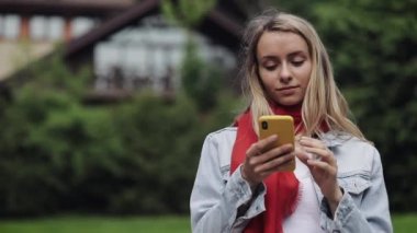 Akıllı telefon kullanarak kırmızı fular giyen genç güzel kadın portresi, telefon kontrol ve bir parkta iyi haber kutluyor. Bir kır evinin yanında duran kız.