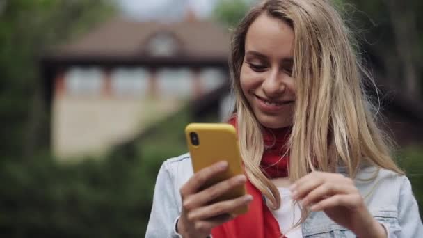 Πορτρέτο της νεαρής όμορφη γυναίκα χρησιμοποιώντας την εφαρμογή στο smartphone, χαμογελά και μηνύματα στο κινητό τηλέφωνο. Γυναίκα φορώντας το κόκκινο κασκόλ στέκεται κοντά σε ένα εξοχικό σπίτι. — Αρχείο Βίντεο