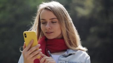 Akıllı telefon kullanarak kırmızı fular giyen genç güzel kadın portresi, telefon kontrol ve bir parkta iyi haber kutluyor. Şehir parkında duran kız.