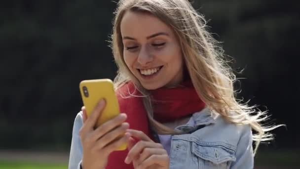 Portré fiatal szép nő segítségével app okostelefon, mosolygós és textil a mobiltelefonon. Nő visel a piros sálat áll a városi parkban.