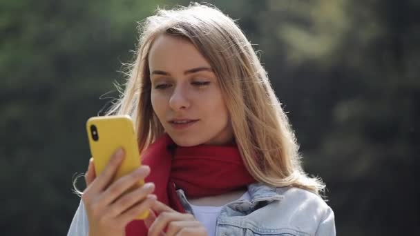 Retrato de una mujer joven y hermosa usando la bufanda roja usando un teléfono inteligente, revisando el teléfono y celebrando las buenas noticias en un parque. Chica de pie en el parque de la ciudad . — Vídeo de stock