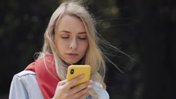 Portrait of young beautiful woman using app on smartphone, wearing in the red scarf standing in the city park. — Stock Video