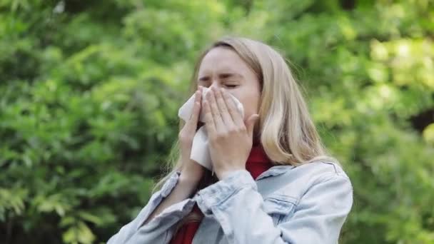 Jonge vrouw met allergie symptoom blazen neus staande in het Park. Ziek meisje niezen en blazen haar neus in weefsel als gevolg van allergie voor boom pollen. — Stockvideo