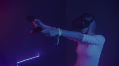 Neon ışıkları arka planda gamepad ile modern sanal gerçeklik kulaklık kullanarak genç çekici kadın. Uzaktan kumandalar, tıklamalar, Vr'da kadın işi, elle gesticulating.