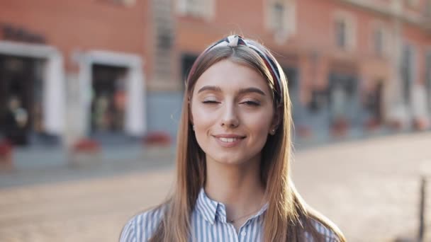 Ritratto di una bella giovane donna che guarda nella macchina fotografica e sorride in piedi sul vecchio sfondo della strada. Ragazza che indossa in abito camicia a righe con fascia . — Video Stock