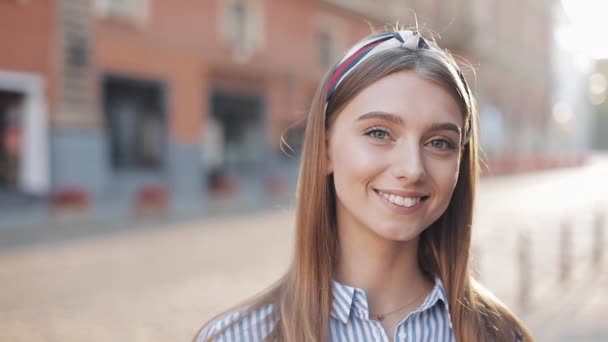 Retrato de una hermosa joven mirando a la cámara y sonriendo de pie sobre el viejo fondo de la calle. Chica usando en vestido de camisa a rayas con diadema . — Vídeos de Stock