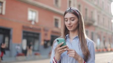 Mavi beyaz çizgili tişört giyen gülümseyen genç bir kadın akıllı telefon kullanarak eski caddede yürüyor. İletişim, sosyal ağlar, online alışveriş kavramı.