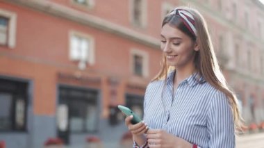 Mavi beyaz çizgili tişört giyen gülümseyen genç bir kadın akıllı telefon kullanarak eski caddede yürüyor. İletişim, sosyal ağlar, online alışveriş kavramı.