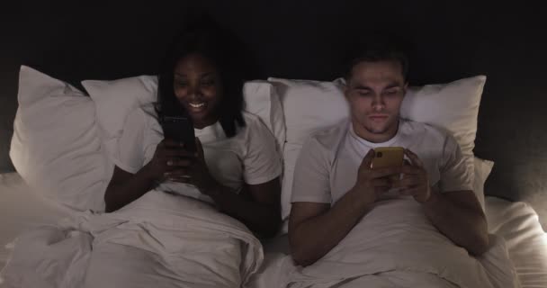 ベッドの中でスマートフォンを持つ幸せな多民族のカップル。人、技術、インターネット、コミュニケーションの概念. — ストック動画