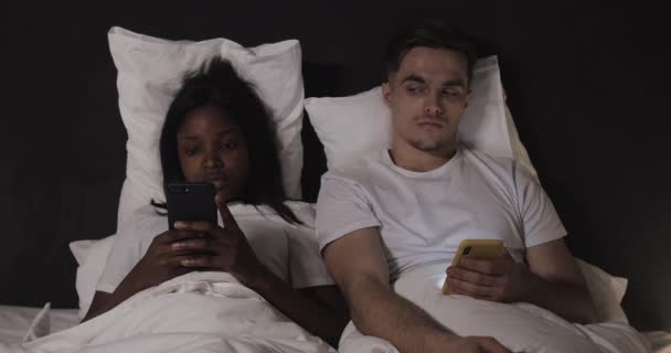 Multi-etnisch koppel met behulp van smartphones liggend in het bed 's nachts. Man Peeks op zijn vrouw s telefoon, ze keert zich af van hem. Spionage, complexe relaties. Geen vertrouwen. — Stockvideo