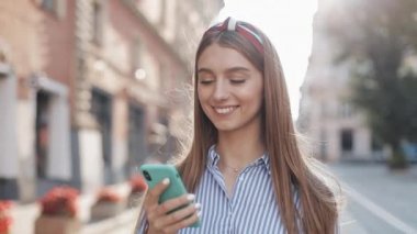 Mavi beyaz çizgili tişört giyen gülümseyen genç bir kadın akıllı telefon kullanarak eski caddede yürüyor. İletişim, sosyal ağlar, online alışveriş kavramı.